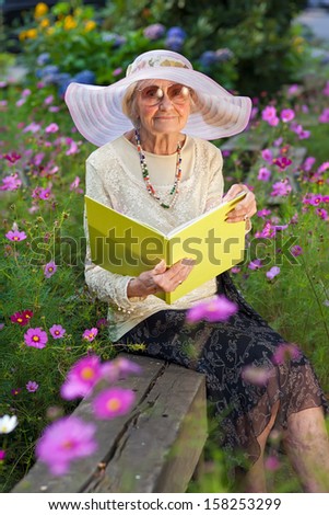 Elegant elderly lady reading in the garden sitting on a wooden bench amongst the flower beds in sunglasses and a stylish wide brimmed sunhat