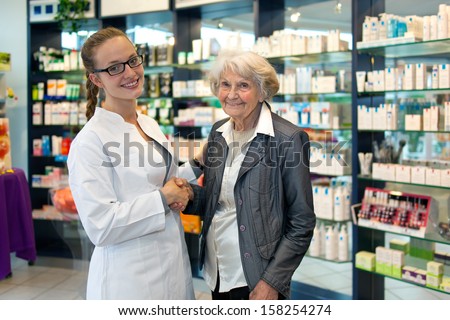 Senior lady with a lovely smile shaking hands with a pretty young female pharmacist as they stand together in the pharmacy discussing the products and medication