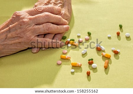 Clasped hands of an elderly lady resting on a table alongside a variety of scattered medication, tablets capsule and pills, prescribed for her health and as diet supplements