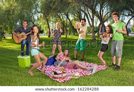 Group of lively happy teenage young friends enjoying a picnic outdoors dancing and singing along to guitar music played by one of the boys