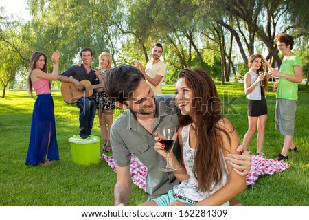 Attractive young teenage couple drinking red wine and enjoying a picnic with friends in the park as they play the guitar and dance