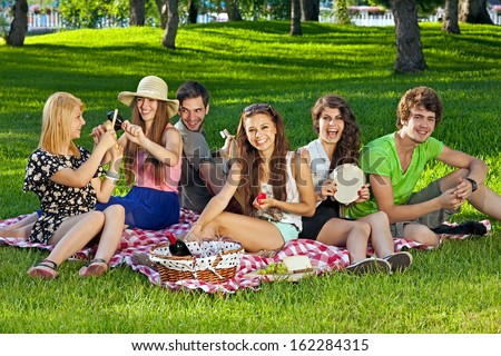 College students enjoying a picnic in the park sitting grouped together on rug on the grass laughing and joking amongst themselves