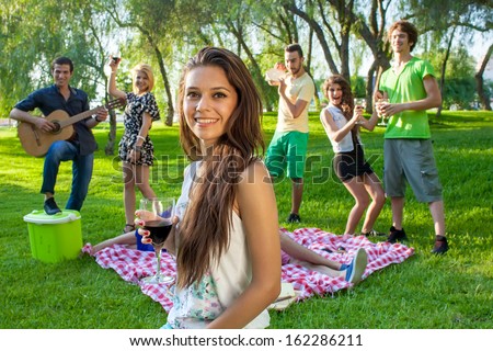 Group of young college friends partying in the park with a pretty young teenage girl in the foreground smiling at the camera and holding a glass of red wine
