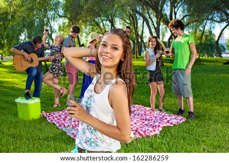 Young teenage girl partying with friends smiling and holding a glass of red wine as her college classmates picnic and play guitar in the background