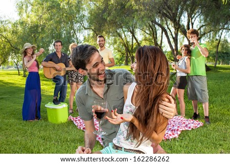 Happy romantic young teenage couple in an affectionate embrace enjoying a good laugh together in the park watched by a group of their college friends as they relax on their summer vacation