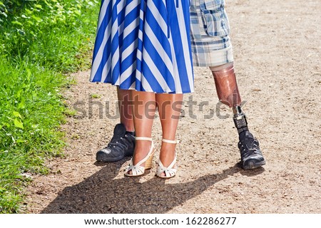 Close up view of the legs of a disabled man wearing a prosthetic leg following a limb amputation standing with a stylish woman in a dress on a gravel path