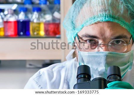 Male scientist working in a laboratory wearing glasses, a surgical cap and mask looking up over the ocular of his microscope at the camera, close up of his head