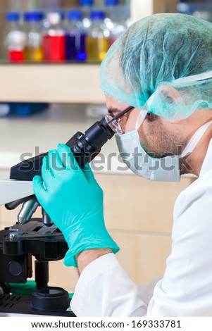 Close up of a male laboratory technician in a surgical cap and mask and wearing glasses looking down a microscope
