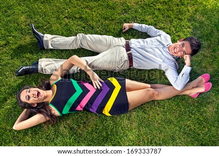 Vivacious teenage couple lying on their backs on lush green grass head to toe resting their arms on each others legs, overhead view of a stylish fashionable young man and woman