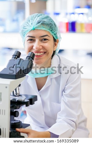 Happy female laboratory technician sitting at her microscope in a surgical cap and mask looking up to smile warmly at the camera