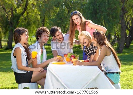 Group of happy attractive young teenagers enjoying their summer vacation sitting down at a table for a picnic together in a lush green park.