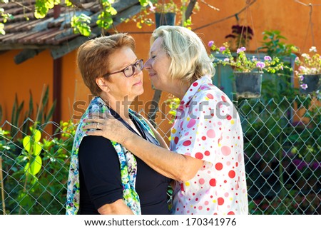 Elderly ladies greeting each other with a kiss on the cheek, leaning closing to whisper a secret or share the latest gossip as they stand outdoors in the garden.