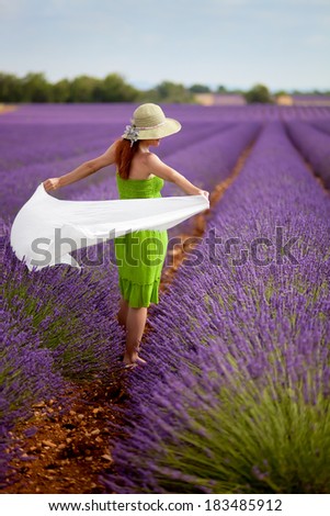 Attractive woman in green dress in lavender field holding long white scarf. Wearing light green hat, decorated with lavender flowers. Blue sky. Summer concept.