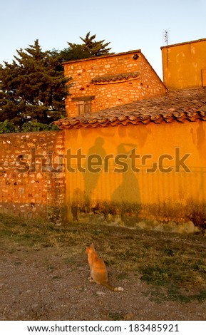 Lone ginger cat sitting in the glow of the late evening sun watching the shadows of a man and woman falling onto an old rural stone wall in Provence, France.