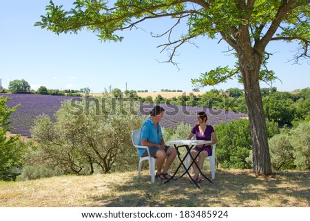 Man and woman next to their tent sitting on camping chairs at table under tree. In background lavender field in Provence, France.