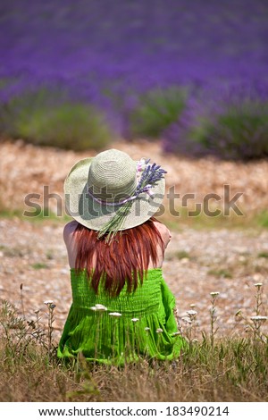 Romantic lady wearing green dress and hat, sitting in front of violet lavender field. Close, back-view. The hat is decorated with lavender twigs.