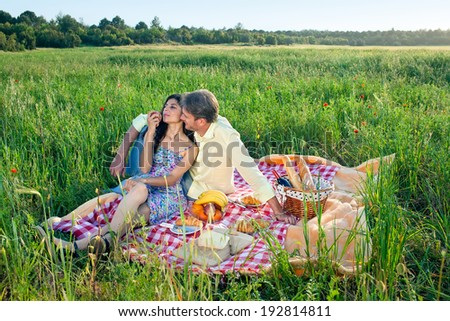 Romantic couple enjoying a summer picnic sitting on a red and white checkered rug in a poppy field eating their meal while leaning intimately against each other
