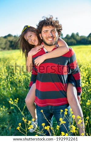 Handsome happy young man giving his pretty girlfriend a piggy back ride through a field of colorful yellow rapeseed flowers on a warm sunny summer day.