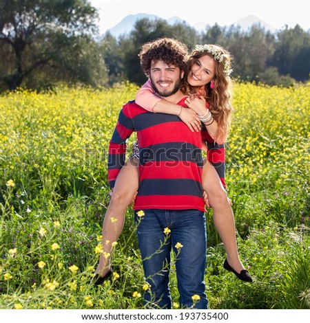 Handsome young man giving his pretty young girlfriend a piggy back ride as they stand smiling at the camera in the middle of a colorful yellow rapeseed field
