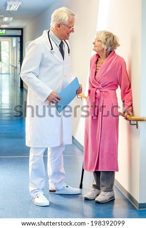 Doctor talking to an elderly woman patient dressed in a pink dressing gown and holding a walking stick in the corridor of the hospital with a file in his hands.