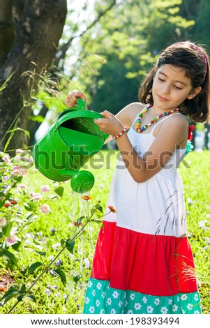 Pretty young girl watering flowers in the garden with a bright green watering can wearing a pretty colorful summer dress and necklace of large beads