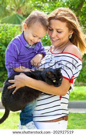 Family portrait with a beautiful young modern mother smiling while holding her adorable little son and a funny black and fat cat, outdoors, in a warm summer day