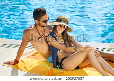 Loving tanned fit healthy young couple relaxing in the sun at the pool in their swimsuits on a colorful yellow towel smiling happily at the camera