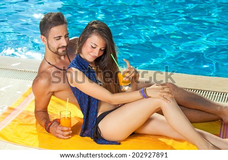 Young couple sunbathing and drinking orange cocktails at the edge of a sparkling blue sunlit pool as they enjoy their summer vacation at a tropical resort