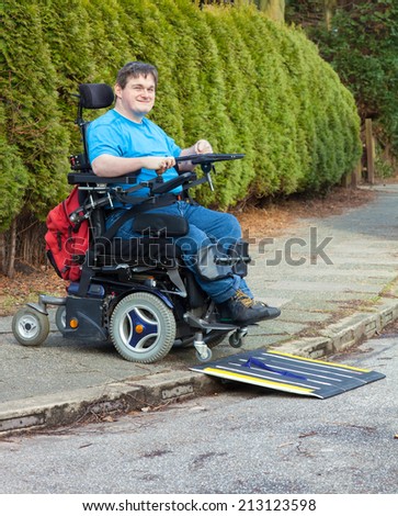 Young man with infantile cerebral palsy caused by birth complications negotiating a mobile ramp on a roadside kerb with his multifunctional wheelchair during a days outing for integration