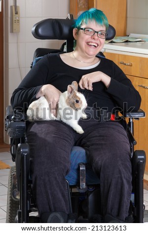 Cheerful young infantile cerebral palsy patient caused by complications at birth sitting in a multifunctional wheelchair stroking a pygmy rabbit as therapy with a beaming smile