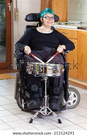 Happy spastic young woman with infantile cerebral palsy due to birth complications confined to a multifunctional wheelchair beating on a drum as part of her therapy smiling at the camera