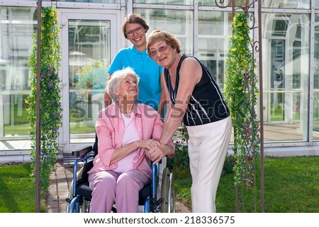 Smiling Care Taker, Looking at Camera, for Old Age Patient on Wheel Chair, visited by an elderly lady. Captured at Grassy Green Garden with Glassy Home Care Building Background.