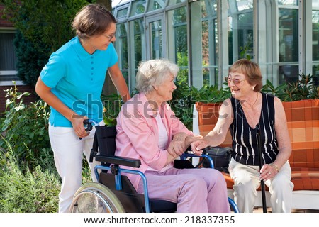Elderly lady sitting in a wheelchair being pushed by a carer pausing to talk to a friend who is sitting on a wooden garden bench