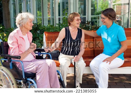 Three women sitting chatting on a garden bench, one old lady sitting in a wheelchair, while one elderly woman has a walking stick.