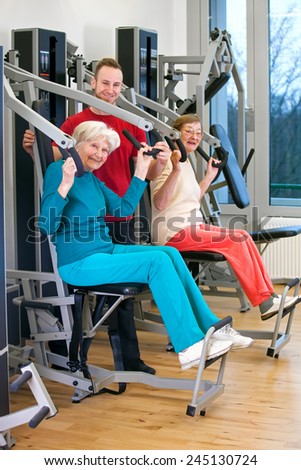 Happy Old Women Exercising Chest Press Workout at the Fitness Gym Assisted by Young Male Instructor.