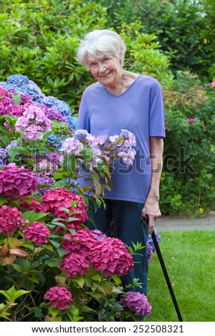 Smiling Old Woman with Cane Posing Beside Pretty Flowers at the Garden While Looking at the Camera.