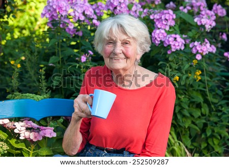 Close up Happy Grandma in Red Shirt Having a Cup of Coffee at the Garden with Flower Plants at the Background.