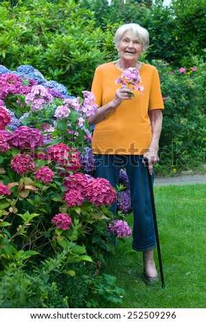 Full Length Shot of Happy Old Woman with Cane Holding Flowers at the Garden While Looking at the Camera.