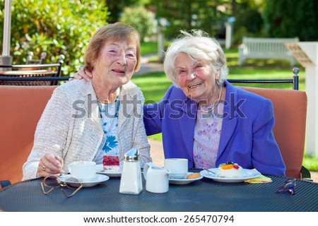 Two Happy Senior Women Relaxing at the Garden Table with Cups of Coffee and Snacks, Looking at the Camera.