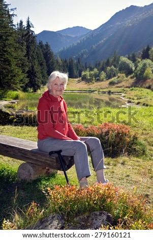 Portrait of Smiling Senior Woman Wearing Red Jacket Sitting on Outdoor Bench near Mountain Valley Lake in Switzerland.