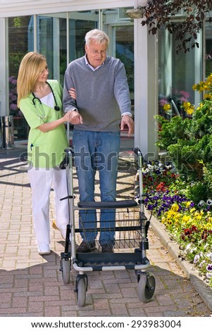 Smiling Blond Nurse Helping Senior Man to Walk with Walker, Steadying Man with Walker Outdoors in front of Building with Flower Gardens.