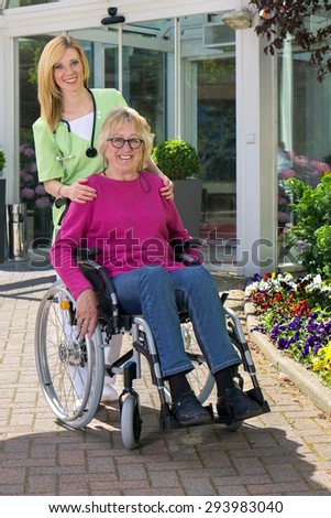 Portrait of Smiling Blond Nurse Standing Behind Senior Woman in Wheelchair Outdoors in front of Building on Sunny Day.