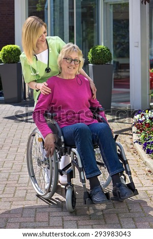 Caring Blond Nurse Standing Behind Senior Woman in Wheelchair with Hand on Shoulder Outdoors in front of Building on Sunny Day.