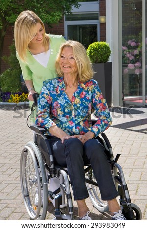 Blond Nurse Standing Behind Senior Woman in Wheelchair with Hand on Shoulder Outdoors in front of Retirement Home on Sunny Day.
