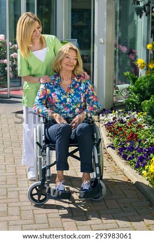Blond Nurse Standing Behind Senior Woman in Wheelchair with Hands on Shoulders Outdoors in front of Building on Sunny Day.
