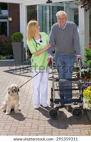 Smiling Blond Nurse Holding onto Arm of Senior Man  Helping Man with Walker Walk Dog on Leash Outdoors in front of Retirement Building on Sunny Day.