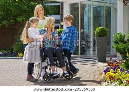 Happy cute caring grandchildren with concerned mother visiting youthful looking grandmother in wheelchair.