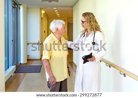 Young Female Doctor with Blood Pressure Apparatus, Talking to an Elderly Woman with Special Needs at the Hospital Corridor.
