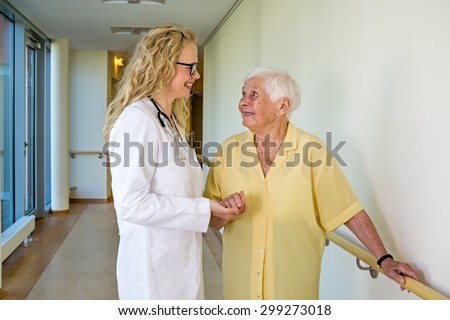 Happy Young Nurse Supporting an Old Woman Walking at the Corridor Inside the Hospital While Holding on the Wall Rail.