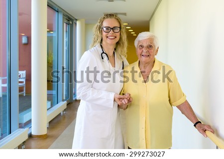 Kind Female Nurse Assisting her Old Woman Patient at the Hallway in the Hospital, Smiling at the Camera